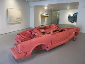 Sylvie Fleury, Skin Crime 6, crashed car, enamel, 31 x 29 x 141 inches, 1997 in foreground of installation view of ‘Shrines to Speed’ at Leila Heller Gallery, June 2016.